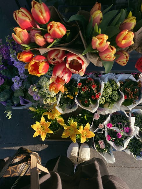 A top-down view of a variety of fresh flower bouquets and potted plants displayed outdoors. Prominent among them are a bouquet of vibrant pink and yellow tulips arranged with long green leaves, and sm