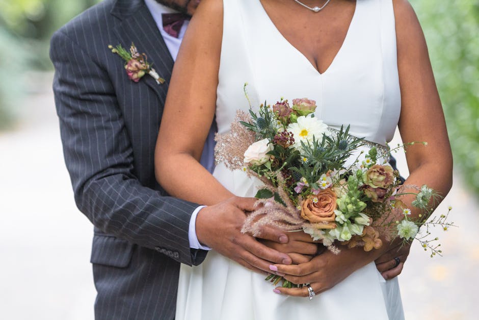 A bride and groom are featured in an outdoor setting, with the groom holding a large, freshly arranged floral bouquet. The bouquet includes soft peach and white roses, light green foliage, delicate da