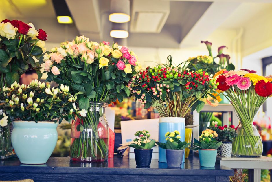 An assortment of fresh flower bouquets and potted plants displayed on a dark wooden table in a well-lit florist shop. The arrangement includes large vases with pastel pink and white roses, a bouquet o